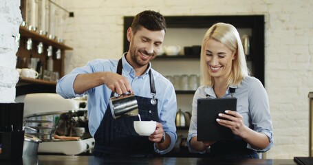 Beautiful blonde woman waitress watching video on the tablet device while attractive waiter pouring milk in the cup of coffee at the bar.