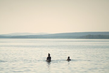 Obraz premium Photo of a summer vacation on the shore of a large reservoir. Silhouettes of a boy and a girl who enter the water, against the backdrop of a picturesque mountain landscape.