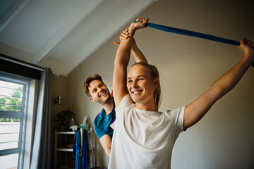 Caucasian female happily performing recovery exercises with rubber band on male physiotherapist bed