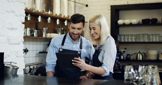 Cheerful good looking waiter and waitress standing at the bar, talking and watching some video on the tablet computer.