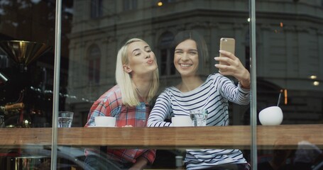 Beautiful two women posing and taking nice selfie photos on the smartphone camera while sitting with coffee in cafe.