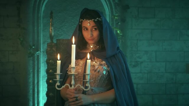 Beautiful young mysterious fairytale woman in halloween costume medieval queen posing model on backdrop stone wall castle. Brunette girl holding candlestick, burning candles. Blue creative cape, hood
