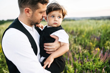 Tender father is carrying toddler in his arms. Dad and little kid in suit vests outdoors