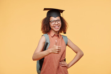 Portrait of cheerful satisfied black graduate student in cap holding hand on hip and showing thumb-up against yellow background