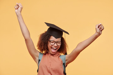 Happy excited black graduate student in eyeglasses and academic cap raising arms and screaming...