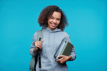 Portrait of positive African-American student girl in hoodie holding stack of books against blue...
