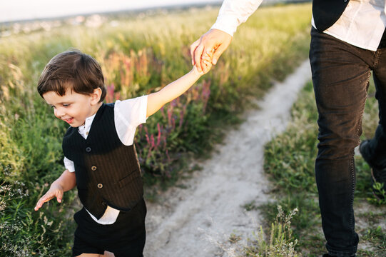 A midshot of a little kid in black vest walking on the field road and laughing. A man's hand is holding his hand. A picture of happiness and trust