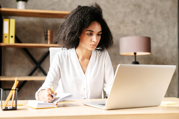 Concentrated african-american young woman in smart casual wear working with papers in modern office. Mixed-race girl making notes on paper documents while working with a laptop