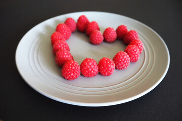 on a grey round plate a heart is laid out with fresh raspberries side view on a black background