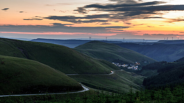 Sunset over the south Wales valleys from the Bwlch mountain.  A road winds around the hillside to the village of Abergwynfi.  The hillside has a wind farm on it. 