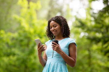 Lovely black lady with takeout coffee browsing social media on cellphone at park