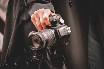 A girl photographer in a black raincoat in rainy weather holds a black umbrella and a SLR camera in her hand.