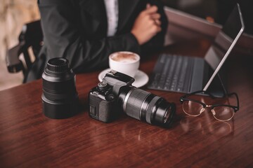A girl photographer in a black raincoat and white shirt in rainy weather is sitting outside at a table after a photo shoot drinking coffee and looking at photos on a tablet. On the table is a camera.