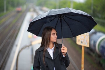 The young girl in a black coat under an black umbrella is walking through the station to the train. Rainy weather .
