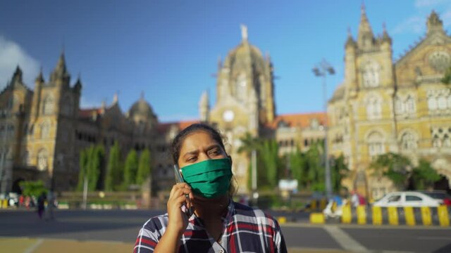 Young Girl Wearing Mask and Talking On Phone Standing Front Of Chhatrapati Shivaji Terminus (CST) formerly Victoria Terminus, is a UNESCO World Heritage Site, Mumbai