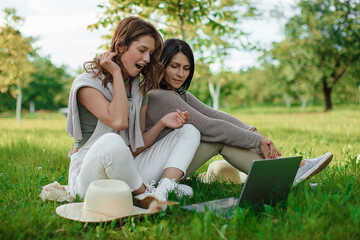 Two beautiful girl in white clothes on a laptop computer outdoors. Lay on the green grass.

