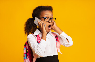 Shocked African Schoolgirl Talking On Cellphone Standing On Yellow Background