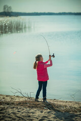 A girl in a pink jacket on the lake shore on an autumn day fishing with a spinning rod