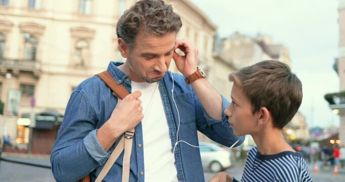 Caucasian cheerful father listening to music with teen son in headphones at street in city. Boy giving headphone to listen favorite son to his dad. Different ages and generations with same tastes.