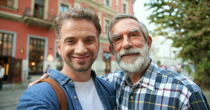 POV of Caucasian happy handsome man taking selfie photo on smartphone camera with senior gray-haired father at street in town. Cheerful old dad and son posing to phone while making photos outside.