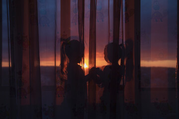 silhouettes of two girls on the windowsill behind the curtain against the sunset sky
