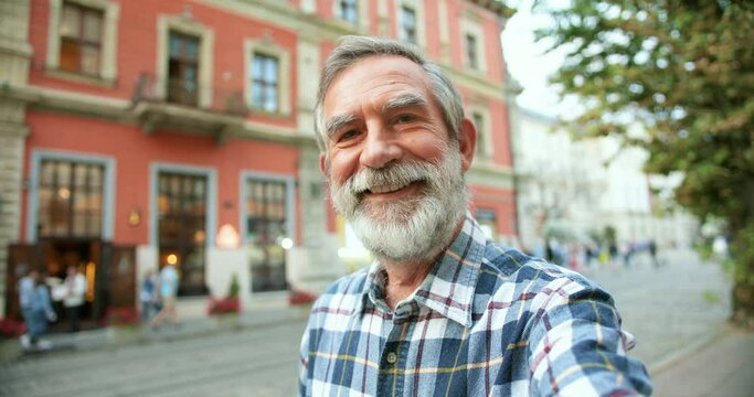 POV of Caucasian happy handsome old man with gray hair taking selfie photo on smartphone camera at street in town. Cheerful senior male with beard posing to phone while making photos outside.