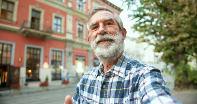 POV of Caucasian happy handsome old man with gray hair taking selfie photo on smartphone camera at street in town. Cheerful senior male with beard giving thumb up while making photos or videochat.