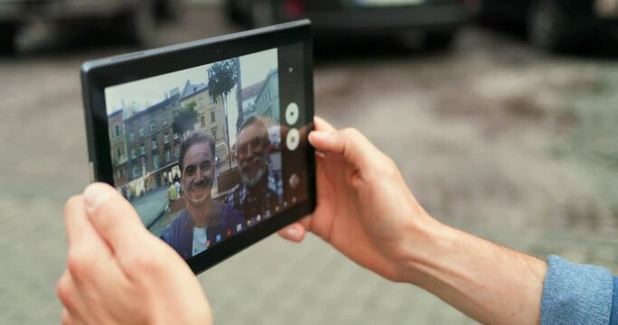 Close up of black tablet device in hands of young and old men. Adult son and senior father taking selfie photo on gadget camera outdoor at street. Screen of computer with photos of dad and son.