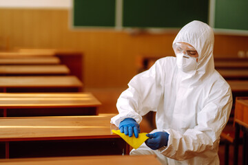 Man in protective hazmat suit washes school desk during coronavirus pandemic. COVID-19.