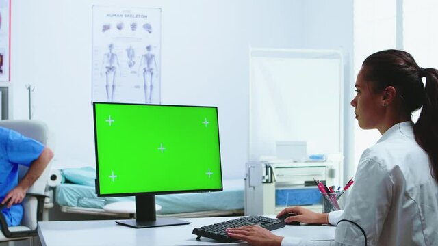 Doctor using computer with green screen display in hospital cabinet and assistant in the background. Medic in white coat working on monitor with chroma key in clinic cabinet to check patient diagnose.