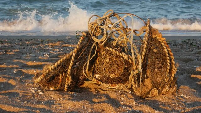 Old used dirty fishing nets with rusty wire, garbage dumped on the sea beach sand. Environmental pollution problem concept. Trashy sandy shore. Moving waves on blue water background. Volunteer concept