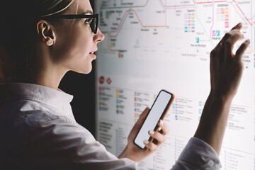 Interactive kiosk with public transport subway map.Female standing at big display with smartphone in hand.Young woman touching with finger screen while using train schedule application on mobile phone