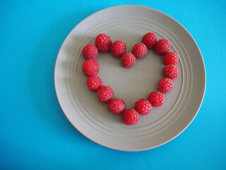 a heart with fresh raspberries is laid out on a grey round plate top view on a blue background