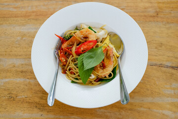 Spicy stir fried flat spaghetti with prawn and holy basil leaves in a white plate on a wooden table with a fork.