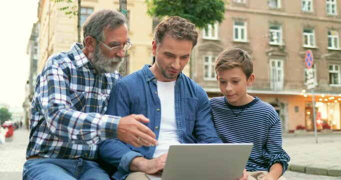 Handsome Caucasian man sitting at bench with old father and teen son and showing something on computer outdoor at street. Males of different ages and generations talking and using laptop in town.