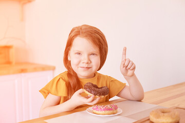 Little girl eating round doughnut. Pointing up. Sweet desert. Unhealthy food. Home lockdown lifestyle. Funny breakfast. Looking not in camera. Choco brown donut. Copyspace. Horizontal banner