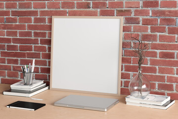 Square poster frame mockup on the wooden table of home studio workspace with red brick wall. Side view, clipping path around poster picture.