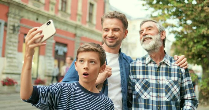 Cute teen Caucasian boy taking selfie photo with father and old grandfather at street in city with smartphone. Senior man with adult son and little grandson posing while making photos on phone.
