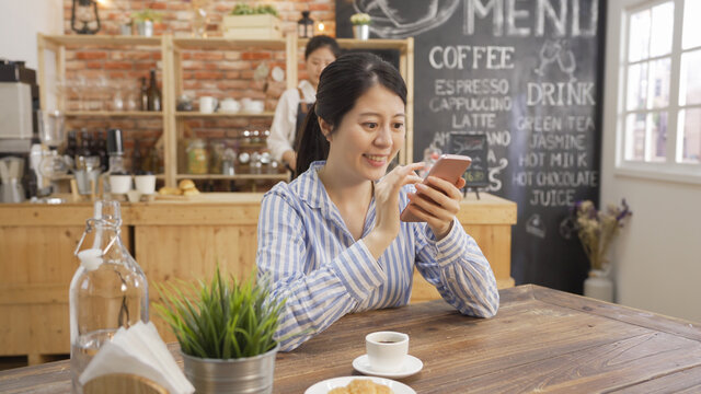 Woman typing writing message on smart phone in modern cafe. young office lady sitting at wooden table with coffee and croissant using mobile phone during breakfast time. bartender working in counter