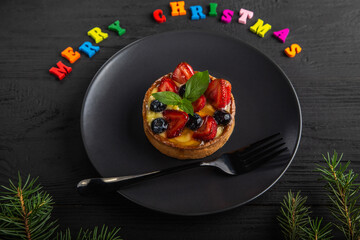 Xmas cake or small tart with berries, cream and mint leaves on a black plate. Fir branches and colorful letters of Merry Christmas on black wooden background