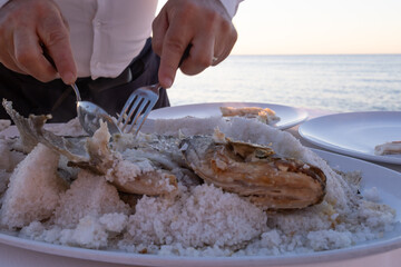 Expensive dinner at sunset on a beach restaurant with amazing sea view. Waiter in white shirt serving the meat of a big sea bass cooked in the oven in a crust of salt.