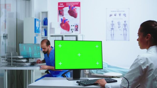 Assistant giving doctor x-ray image while working on computer with green screen monitor in hospital cabinet. Desktop with replaceable screen in medical clinic while doctor is checking patient