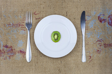 A slice of kiwi on a white plate with a fork and a knife on a tablecloth.