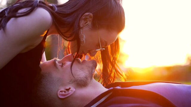 close up of a kiss at sunset. Attractive couple guy lying on the lawn brunette girl with long hair kisses her nose and lips. Stylish fashion hipsters kissing in nature. Man and woman in love