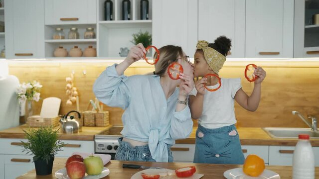 Young mother and African daughter make vegetable salad in kitchen at home. Family is having fun together, covering eyes with red paprika circles looking like eyewear. Smile and kiss playful mood.