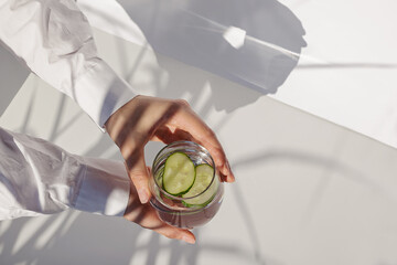 Woman's hands holding glass of water with cucumber on the white table