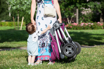Capricious child asks mother to pick up in arms, unrecognizable woman holding baby pushchair in...