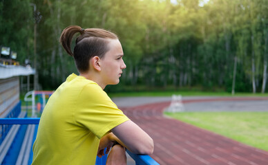 Fototapeta na wymiar teenager boy with topknot haicut. top knot or samurai men hairstyle. teen guy looking at the stadium. back view. serious teenager ina yellow t-shirt. generation z urban style.