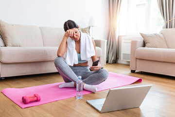 Attractive young woman taking a break while exercising at home. Woman using smart phone on exercise mat in front of her laptop