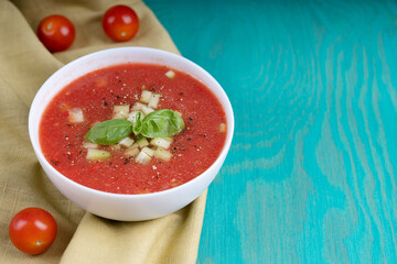 Red gazpacho soup made of red ripe pureed tomatoes and other vegetables decorated with basil leaves and cucumber in white bowl standing on olive towel on blue wooden table. Image with copy space